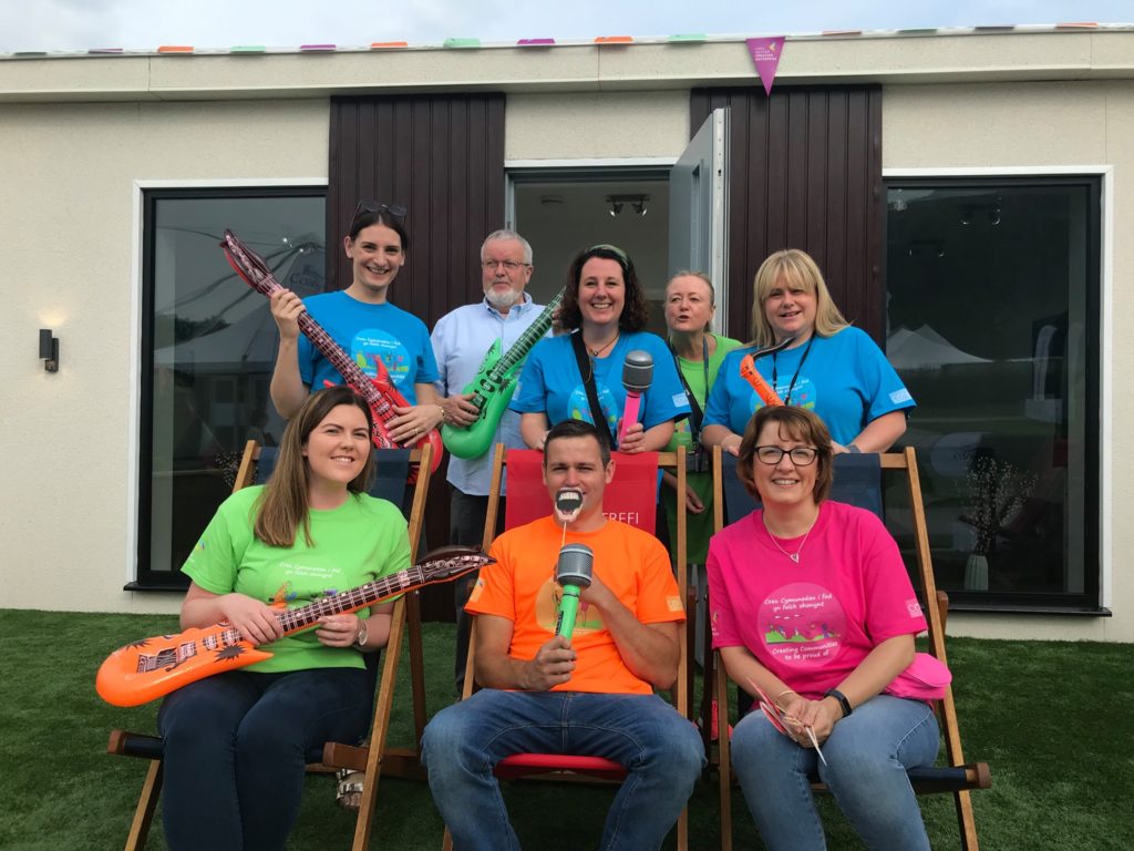 A group of staff in colourful t shirts and inflatable guitars and microphones smile to camera in front of the Haus4One pod