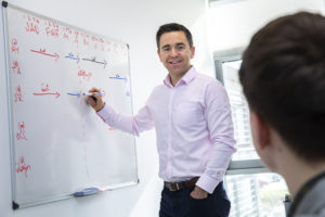 Male staff member smiles to camera and writes on a whiteboard in a meeting